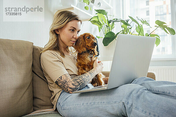 Berufstätige Frau sitzt mit Hund und arbeitet zu Hause am Laptop