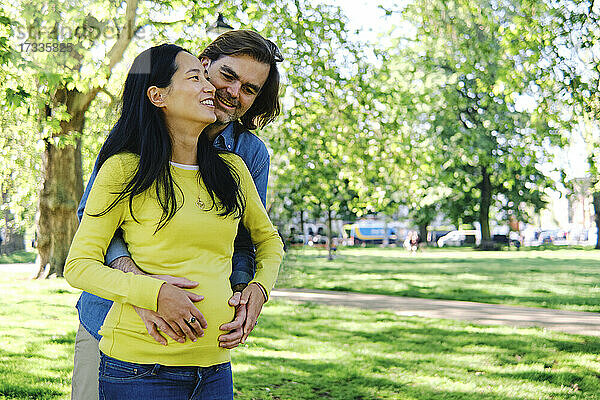 Lächelnder Mann  der seine Frau von hinten umarmt  während er in einem öffentlichen Park steht