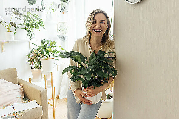Glückliche Frau  die eine Pflanze hält und sich an die Wand im Wohnzimmer lehnt