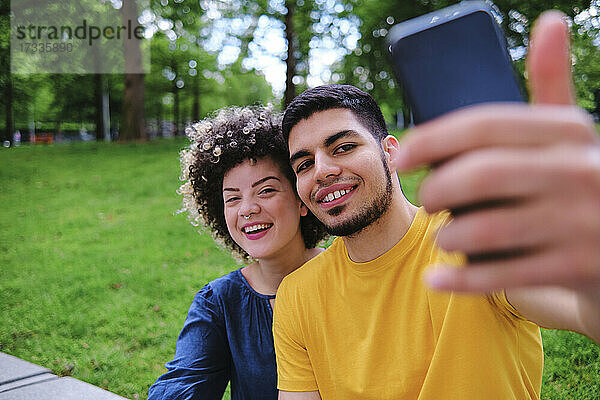 Glückliches junges Paar nimmt Selfie durch Smartphone sitzen im Park