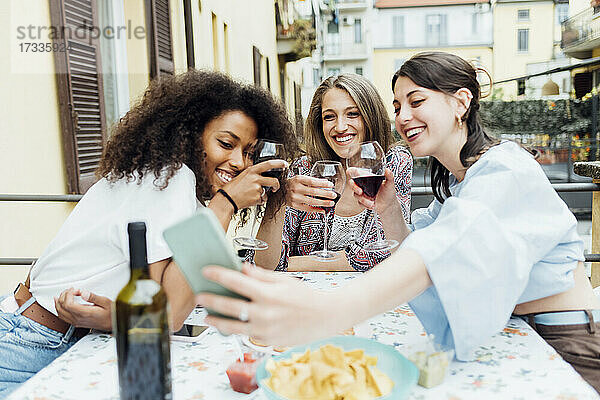 Lächelnde Freunde mit Weingläsern machen ein Selfie mit ihrem Handy auf der Terrasse bei einem geselligen Beisammensein