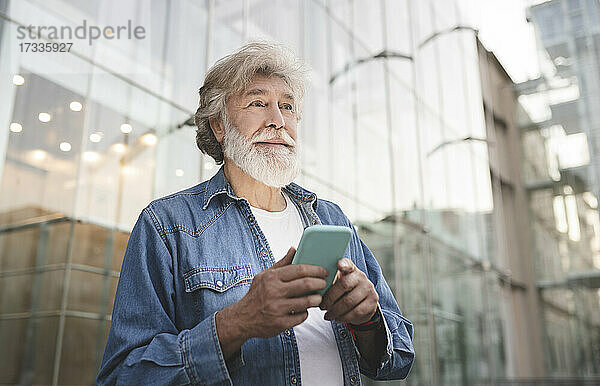 Älterer Mann  der sein Smartphone hält und vor einem Glasgebäude wegschaut