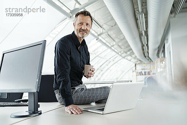 Männlicher Profi mit Kaffeetasse und Laptop im Büro sitzend