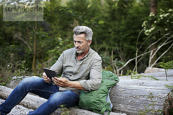 Männlicher Wanderer benutzt ein digitales Tablet  während er auf einem Baumstamm sitzt