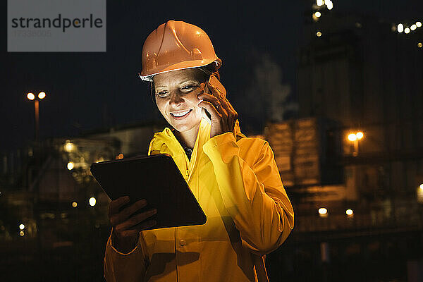 Lächelnde weibliche Fachkraft  die nachts mit einem Smartphone telefoniert und ein digitales Tablet in der Hand hält