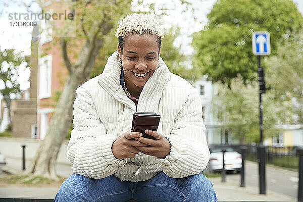 Glückliche erwachsene Frau in weißer Jacke  die ein Smartphone benutzt und im Park sitzt