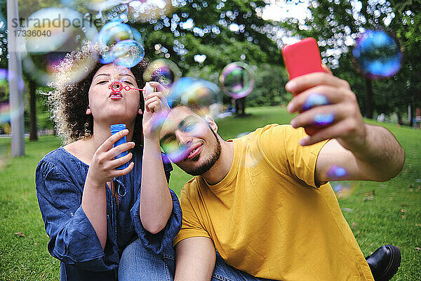 Junger Mann  der ein Selfie mit seiner Freundin macht  die im Park sitzt und Seifenblasen bläst