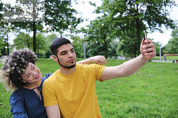 Junger Mann nimmt Selfie mit Freundin durch Smartphone beim Sitzen im Park