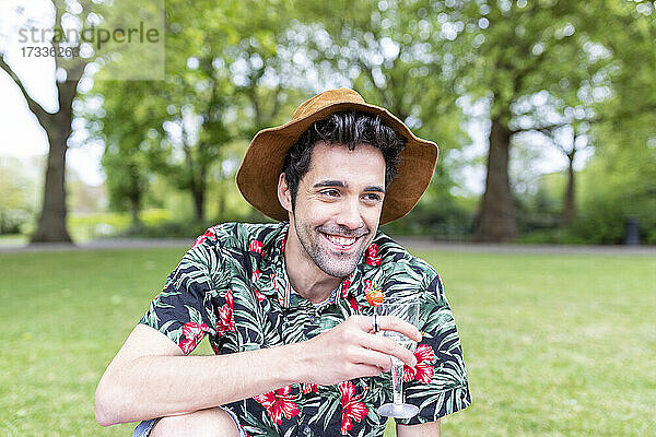 Lächelnder Mann mit Hut  der ein Getränk hält  während er im Park sitzt