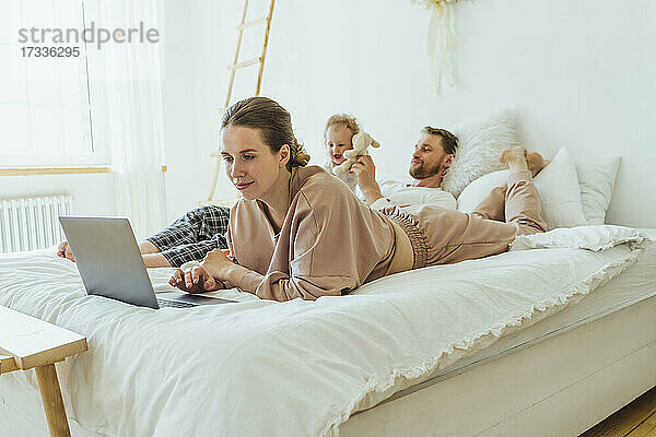 Frau benutzt Laptop  während der Mann mit seiner Tochter im Schlafzimmer zu Hause spielt