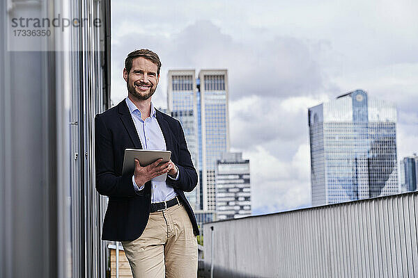 Lächelnder Geschäftsmann mit digitalem Tablet  der sich an eine Gebäudeterrasse lehnt