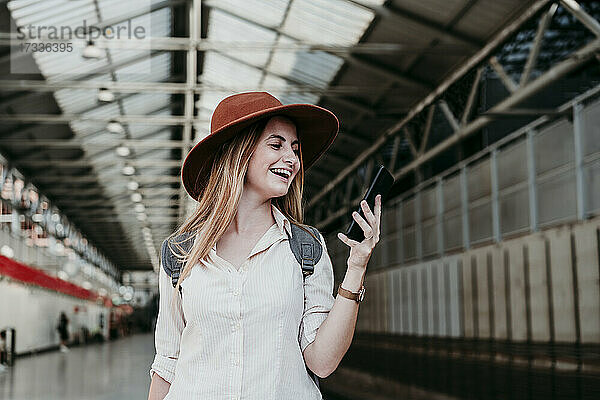 Fröhliche junge Frau mit Hut und Smartphone am Bahnhof