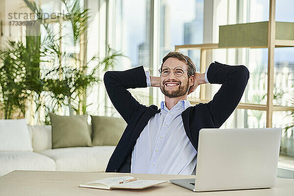 Glücklicher  entspannter männlicher Freiberufler  der mit den Händen hinter dem Kopf im Heimbüro sitzt