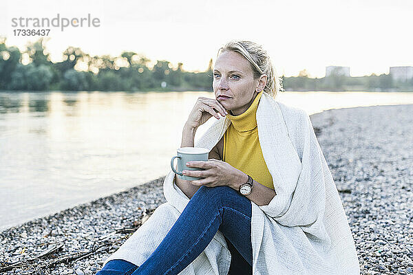 Nachdenkliche reife Frau hält eine Kaffeetasse  während sie am Flussufer sitzt