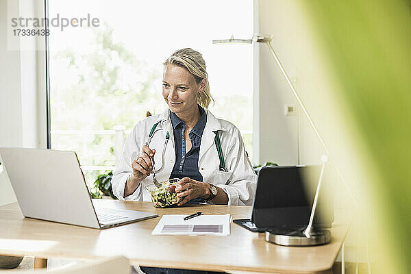 Lächelnde Ärztin isst Essen und schaut auf einen Laptop im Büro