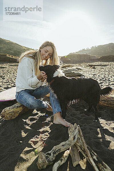 Junge blonde Frau  die ihren Hund streichelt  während sie auf einem Baumstamm am Strand sitzt