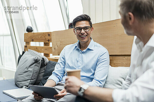 Lächelnde männliche Fachkraft mit digitalem Tablet  die einen Kollegen im Büro betrachtet