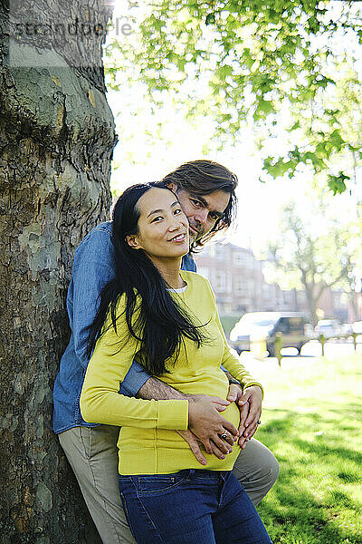 Schwangere Frau lächelt  während ihr Mann sie von hinten im öffentlichen Park umarmt