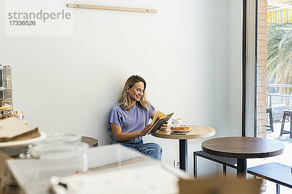 Lächelnde junge Frau liest ein Buch  während sie an einem Tisch in einem Café sitzt