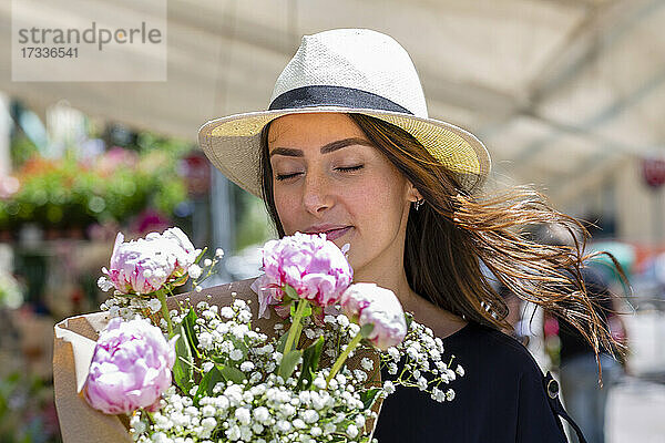 Frau mit Sonnenhut riecht an Pfingstrosen Blumenstrauß in der Nähe von Blumenladen