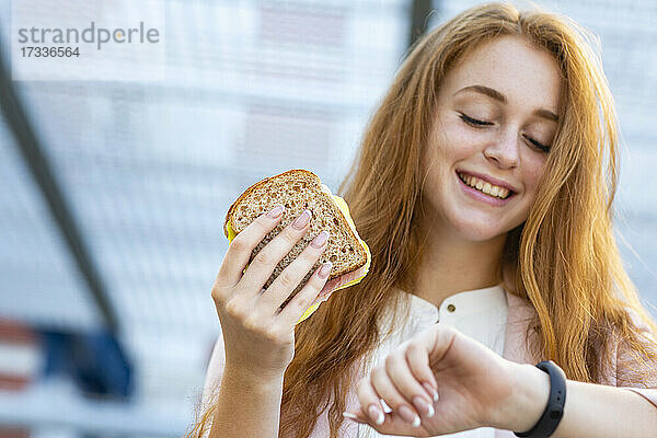 Lächelnde weibliche Fachkraft  die die Zeit überprüft  während sie ein Sandwich am Bahnhof isst