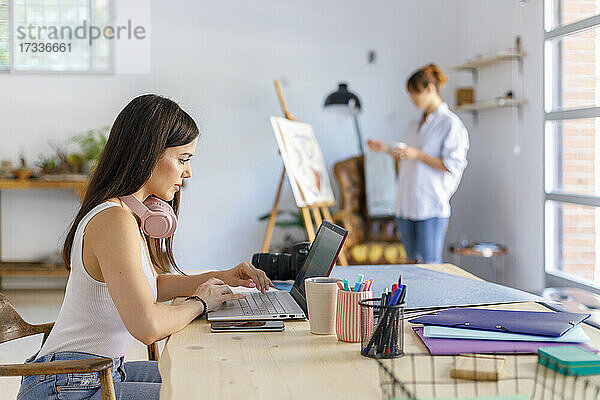 Fotografin benutzt Laptop  während Künstlerin im Atelier malt