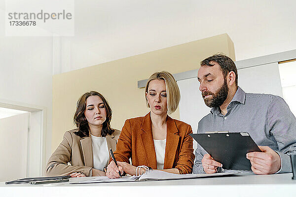 Männliche und weibliche Kollegen  die eine reife Geschäftsfrau beim Schreiben beobachten  während sie während einer Besprechung im Büro diskutieren
