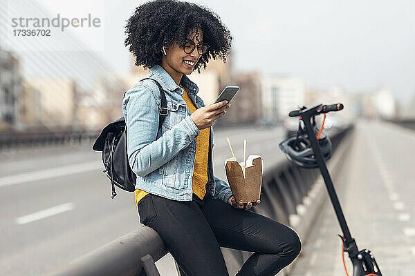 Lächelnde Frau  die ein Smartphone benutzt und Essen in der Hand hält  während sie sich auf ein Geländer stützt