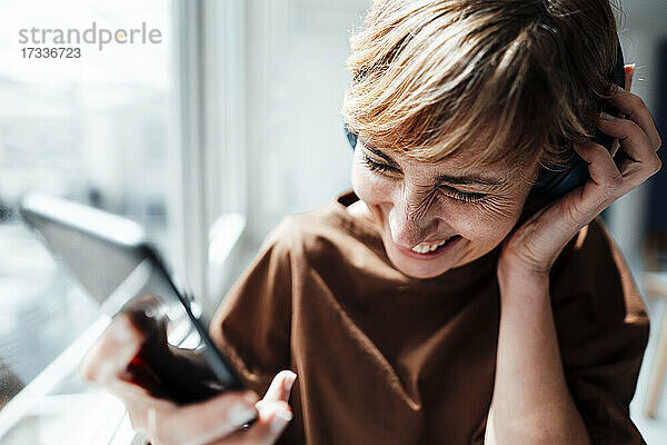 Fröhliche Geschäftsfrau mit Mobiltelefon hört Musik über Kopfhörer im Büro