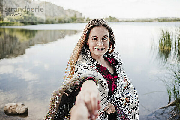 Glückliche junge Frau in eine Decke gewickelt  die die Hand einer Freundin am Seeufer hält