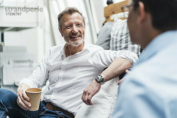 Lächelnder männlicher Fachmann  der einen Einwegbecher hält  während er mit einem Kollegen im Büro sitzt