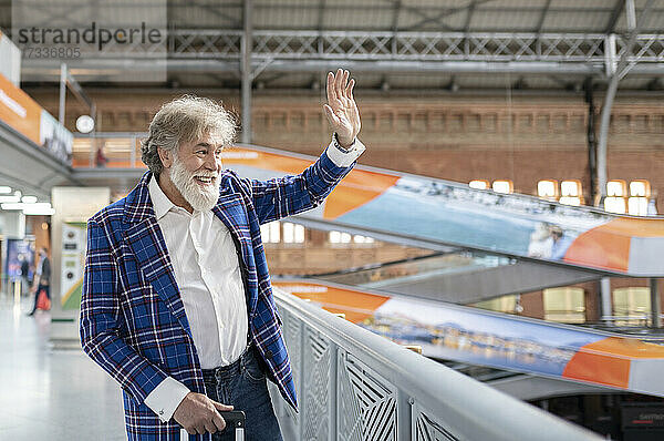 Älterer Mann winkt mit der Hand  während er am Geländer des Bahnhofs steht