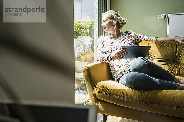 Frau mit digitalem Tablet schaut durch das Fenster  während sie auf dem Sofa im Wohnzimmer sitzt