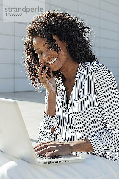 Lächelnde junge Geschäftsfrau  die mit ihrem Laptop vor einem Bürogebäude sitzt und mit einem Mobiltelefon spricht