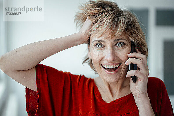 Geschäftsfrau mit Hand im Haar  die im Büro mit einem Mobiltelefon spricht