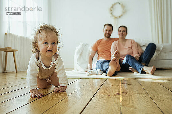 Niedliches Mädchen krabbelt  während Mutter und Vater zu Hause auf dem Boden sitzen
