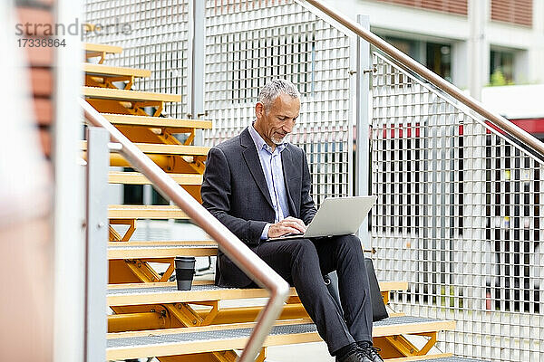 Männlicher Berufstätiger in Geschäftskleidung  der auf einer Treppe sitzend am Laptop arbeitet