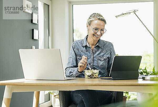 Lächelnde Geschäftsfrau beim Essen  während sie zu Hause ein digitales Tablet benutzt