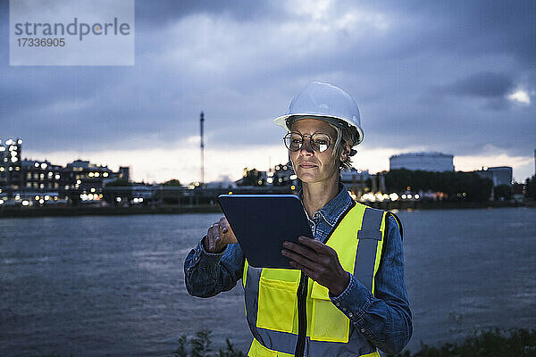 Weibliche Fachkraft in reflektierender Kleidung mit digitalem Tablet am Fluss in der Dämmerung