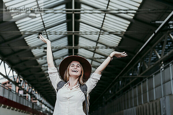 Fröhliche junge Frau  die mit erhobenen Händen am Bahnhof ihren Urlaub genießt