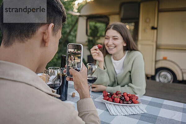 Junger Mann fotografiert Freundin hält Erdbeere durch Smartphone sitzen am Tisch