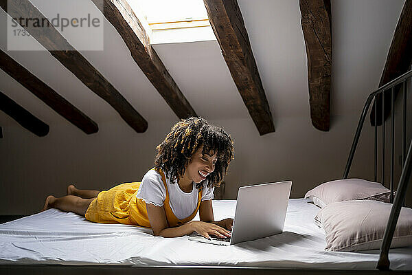 Lächelnde junge Frau  die einen Laptop benutzt  während sie auf einem Bett auf dem Dachboden liegt