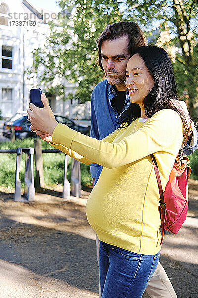 Lächelnde schwangere Frau nimmt Selfie durch Handy mit Ehemann im öffentlichen Park