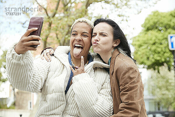 Frau streckt ihre Zunge heraus  während ihre Freundin ein Selfie mit dem Handy macht