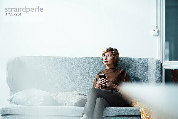 Nachdenkliche Geschäftsfrau mit Mobiltelefon auf dem Sofa sitzend