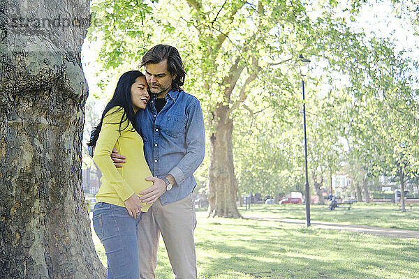 Frau umarmt ihren Mann vor einem Baumstamm im Park