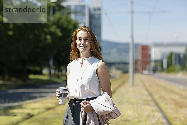 Lächelnde Freiberuflerin mit Einweg-Kaffeebecher an einem Eisenbahnregal stehend
