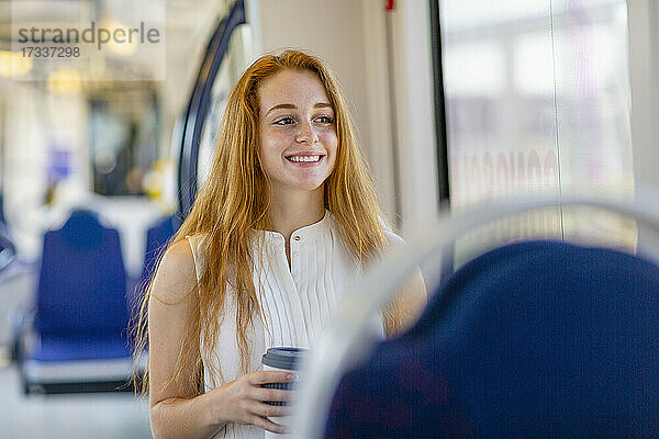 Lächelnde Freiberuflerin mit langen Haaren  die eine Einweg-Kaffeetasse hält und im Zug wegschaut