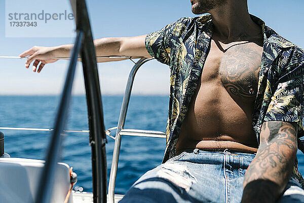 Hipster-Mann mit aufgeknöpftem Hemd auf einer Yacht sitzend an einem sonnigen Tag