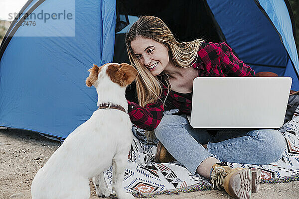 Fröhlicher junger digitaler Nomade streichelt Hund  während er mit Laptop vor dem Zelt sitzt